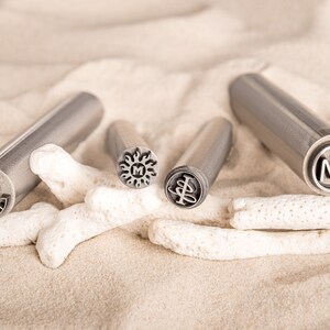 May include: Four metal stamps with different designs, including a sun with an "M" inside, a tree, a letter "Z" and the letters "MA". The stamps are on a white sandy surface with white coral.