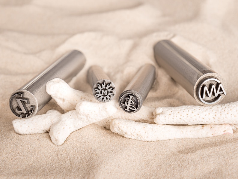 May include: Four metal stamps with different designs, including a sun with an "M" inside, a tree, a letter "Z" and the letters "MA". The stamps are on a white sandy surface with white coral.