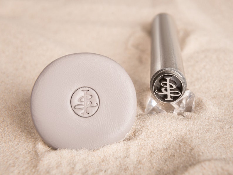 May include: A close-up of a silver metal stamp with a monogram design. The stamp is on a white surface with sand.