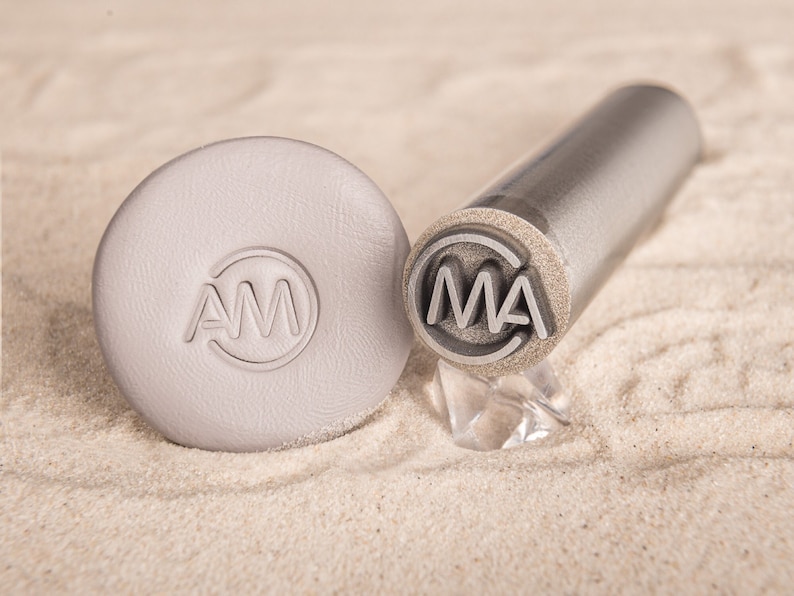 May include: A close-up of two metal stamps with the letters "AM" and "MA" engraved on them. The stamps are on a light brown sandy surface.