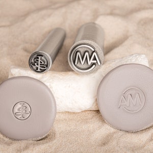 May include: Three metal stamps with different designs. The stamps are on a light brown surface. The designs are "IB", "MA", and "AM".