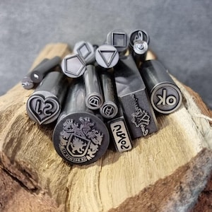 May include: A collection of metal stamps with various designs, including geometric shapes, letters, and a crest with a lion. The stamps are arranged on a piece of wood.