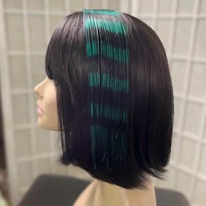 May include: A black bob wig with blunt bangs and teal highlights. The wig is styled with a side part and the teal highlights are concentrated near the front of the hair. The wig is on a mannequin head.