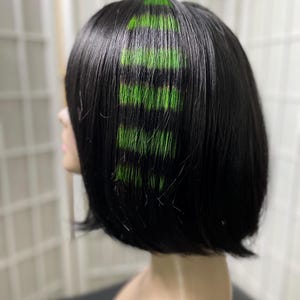 May include: A black bob wig with vibrant green highlights. The wig is styled with a blunt cut, and the green streaks are strategically placed throughout the hair, creating a bold and modern look. The wig is displayed on a mannequin head.