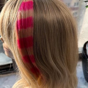 May include: A wig with light brown hair and a vibrant pink streak. The pink streak is composed of several horizontal bands of color, adding a bold contrast to the natural hair color. The wig is styled with soft waves.