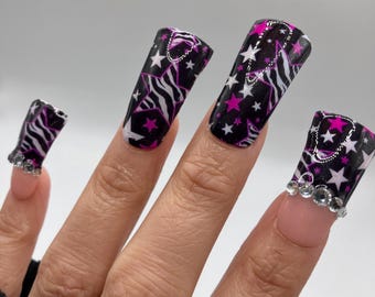 May include: Close-up of a hand wearing long, black and white zebra print press-on nails with pink stars and rhinestones.