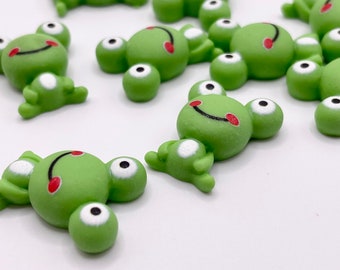 Puede incluir: Un grupo de figuritas de ranas verdes con ojos negros y sonrisas rojas. Las ranas están hechas de un material liso y gomoso y miden aproximadamente 2,5 cm.