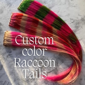 May include: Three custom color raccoon tails in shades of pink, green, and brown. The text "Custom color Raccoon Tails" is displayed in white. The hair extensions are arranged in a curved pattern.