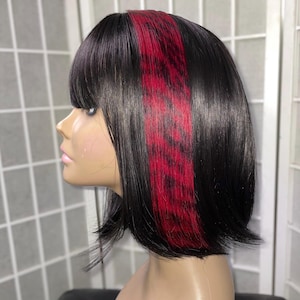 May include: A black bob wig with a bold red highlight. The wig features a blunt cut with bangs and a side profile view. The red highlight is a striking vertical stripe, adding a pop of color to the otherwise dark hair.