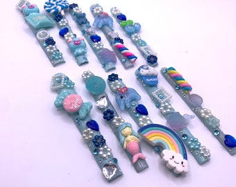 Può includere: Un set di 12 mollette per capelli con glitter blu e vari ciondoli color pastello, tra cui un unicorno, una sirena, un arcobaleno, un cupcake, un lecca-lecca e una ciambella.