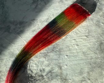 Könnte beinhalten: Eine lebendige, mehrfarbige Haarverlängerung, die von Rot über Grün zu Schwarz übergeht. Die Verlängerung ist eine Mischung aus gefärbten Strähnen, die einen Farbverlauf erzeugt. Das Haar ist gebündelt, wodurch die Farbvariationen zur Geltung kommen.