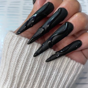 Op de afbeelding: Close-up van een hand met lange, zwarte, stiletto nagels met een druppelend, abstract ontwerp. De nagels zijn geverfd in mat zwart met een glanzend zwart druppelend ontwerp.
