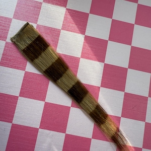 May include: A hair extension piece with alternating bands of light brown and dark brown. The hair extension is rectangular at the top and tapers to a point. The background is a pink and white checkered pattern.