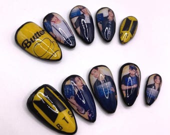 Puede incluir: Conjunto de diez uñas postizas con un diseño amarillo y negro. Las uñas presentan imágenes de una banda de K-pop y las palabras "Butter" y "BTS".