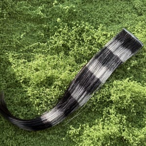 May include: A single hair extension weft featuring alternating horizontal stripes of solid black and bright white. The hair is straight, smooth, and attached to a reinforced top band, creating a bold, high-contrast pattern throughout the length.