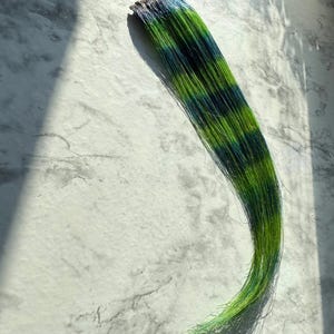 May include: A bundle of green and blue hair extensions. The hair is a mix of vibrant green and deep blue, creating a striking color contrast. The extensions are arranged in a flowing, curved shape, with the ends slightly curled.