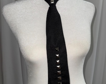 May include: A black necktie featuring a vertical row of silver-toned pyramid studs running down the center of the blade, with two additional matching studs positioned at the knot. The tie is made of a solid black fabric and has a pointed tip.