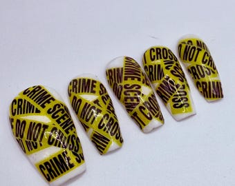 May include: Five white artificial nails with a yellow and black crime scene tape design. The tape reads "CRIME SCENE DO NOT CROSS" and "CRIMINAL". The nails are in a coffin shape.