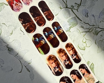 May include: A strip of nail art stickers depicting characters from the film Labyrinth. The stickers show images of the Goblin King and Sarah, in different poses and costumes. The stickers are oval and have a shiny finish.