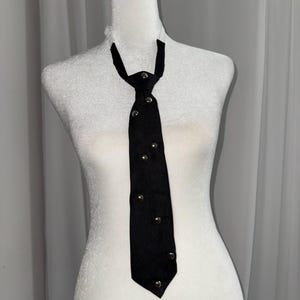 May include: A black necktie featuring a series of small, round, silver-toned metal studs scattered across the front surface. The tie is secured with a white collar band at the top, presenting a clean, structured appearance against the mannequin.