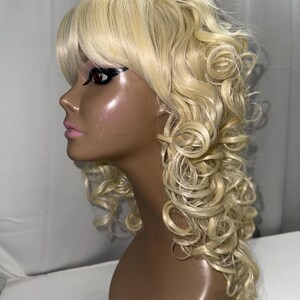 May include: A long, platinum blonde wig with soft, cascading curls and straight-cut bangs. The wig is displayed on a dark-toned mannequin head, showcasing the layered texture and the light, bright color of the synthetic hair fibers throughout.