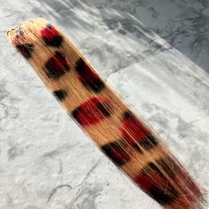 May include: A hair extension piece with a leopard print design. The extension features a light brown base with red and black spots. The hair piece is rectangular and appears to be made of synthetic hair.