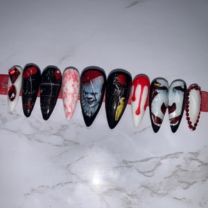 May include: A set of ten long, pointed false nails with a Halloween theme. Designs include a white nail with red gems, black nails with a red balloon, and nails featuring a clown and blood drips. The nails are arranged on a red strip.