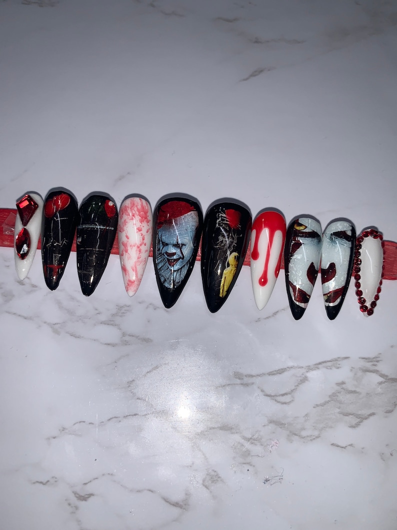 May include: A set of ten long, pointed false nails with a Halloween theme. Designs include a white nail with red gems, black nails with a red balloon, and nails featuring a clown and blood drips. The nails are arranged on a red strip.