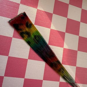May include: A colorful hair extension with a rainbow gradient, transitioning from red to green and blue, with black leopard print spots. The hairpiece is set against a pink and white checkered background.