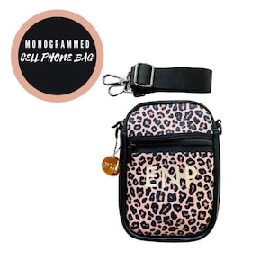 Peut inclure: Un sac de téléphone portable en imprimé léopard rose et noir avec une bandoulière noire réglable et un charme doré avec le mot "Prada" dessus. Le sac a une poche zippée et les initiales "ENP" sont brodées à l'avant en or.