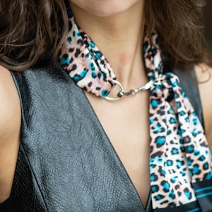 May include: A woman with long, wavy brown hair is wearing a black leather sleeveless top with a blue and brown animal print scarf tied around her neck.