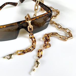 Puede incluir: Gafas de sol carey con una cadena dorada y una cadena marrón y dorada. Las gafas de sol tienen forma rectangular. Las cadenas están conectadas a las gafas de sol y tienen un cierre de mosquetón.