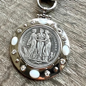K&ouml;nnte beinhalten: Silberfarbener Anh&auml;nger mit detailliertem M&uuml;nzdesign mit drei Figuren und den Worten "FOEDUS INVIOLABILE". Der Anh&auml;nger ist mit klaren Kristallen und Perlmutt-Inlays verziert, die an einem Silberring befestigt sind.