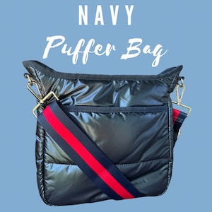 May include: A navy blue puffer bag with a red and black adjustable strap. The bag has a zipper closure and a front pocket.