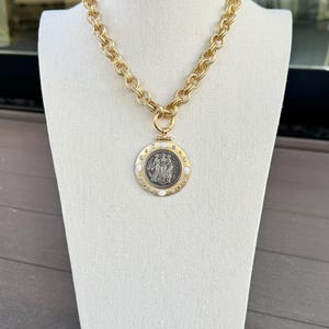 K&ouml;nnte beinhalten: Goldfarbene Kette mit einem gro&szlig;en, kreisf&ouml;rmigen Anh&auml;nger. Der Anh&auml;nger zeigt ein detailliertes, silberfarbenes Relief von Figuren und ist von wei&szlig;en Akzenten umgeben. Die Halskette wird auf einer wei&szlig;en Schaufensterpuppe pr&auml;sentiert.