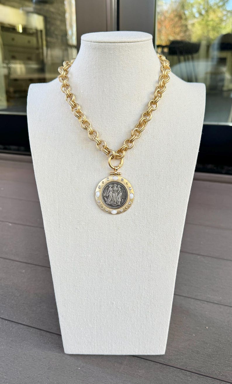 K&ouml;nnte beinhalten: Goldfarbene Kette mit einem gro&szlig;en, kreisf&ouml;rmigen Anh&auml;nger. Der Anh&auml;nger zeigt ein detailliertes, silberfarbenes Relief von Figuren und ist von wei&szlig;en Akzenten umgeben. Die Halskette wird auf einer wei&szlig;en Schaufensterpuppe pr&auml;sentiert.