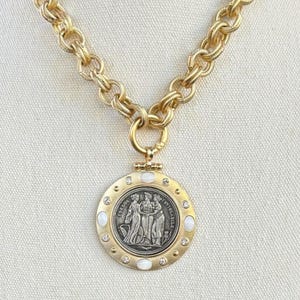 K&ouml;nnte beinhalten: Goldfarbene Halskette mit einem gro&szlig;en, runden Anh&auml;nger mit einem detaillierten Relief von Figuren. Der Anh&auml;nger ist mit kleinen, klaren Kristallen und wei&szlig;en Steinen verziert. Die Kette hat ein klobiges, strukturiertes Design.