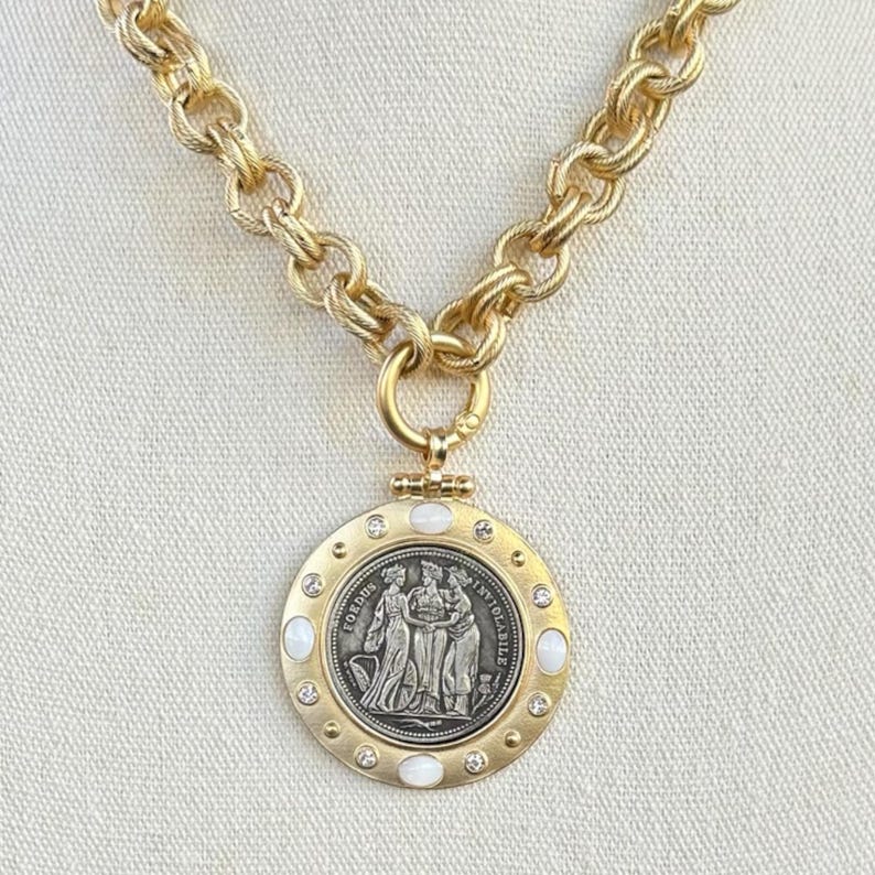 K&ouml;nnte beinhalten: Goldfarbene Halskette mit einem gro&szlig;en, runden Anh&auml;nger mit einem detaillierten Relief von Figuren. Der Anh&auml;nger ist mit kleinen, klaren Kristallen und wei&szlig;en Steinen verziert. Die Kette hat ein klobiges, strukturiertes Design.