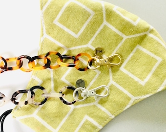 Op de afbeelding: Een groen en wit geometrisch patroon stoffen mondkapje met twee verschillende ketting-stijl maskerstrikken. Eén strik is gemaakt van bruin en zwart schildpad kunststof en de andere is gemaakt van wit en bruin kunststof. Beide strikken hebben een gouden metalen sluiting met een klein gouden label dat "pru" zegt.