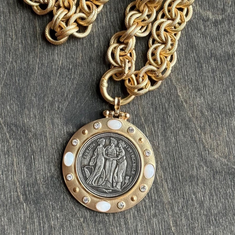 K&ouml;nnte beinhalten: Goldfarbene Halskette mit einem gro&szlig;en, runden Anh&auml;nger. Der Anh&auml;nger zeigt ein silberfarbenes M&uuml;nzdesign mit drei Figuren und den Worten "FOEDUS" und "INVIOLABILE". Der Anh&auml;nger ist mit kleinen Kristallen und wei&szlig;en ovalen Einlagen verziert. Die Kette ist dick und strukturiert.