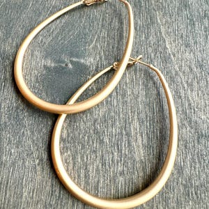 May include: A pair of large, gold-toned oval hoop earrings. The earrings are made of a smooth, shiny metal.