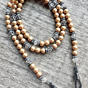 Könnte beinhalten: Ein goldfarbenes Perlen-Lanyard mit klaren und schwarzen Akzenten. Das Lanyard hat kleine, runde Perlen und klare, kristallartige Perlen. Das Lanyard hat zwei schwarze Metall-Drehhaken zum Befestigen an Gegenständen.