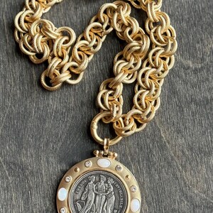 K&ouml;nnte beinhalten: Goldfarbene Halskette mit einem gro&szlig;en, kreisf&ouml;rmigen Anh&auml;nger, der ein detailliertes Relief von Figuren und die Inschrift "FOEDUS INVIOLABILE" aufweist. Der Anh&auml;nger ist mit klaren Kristallen und wei&szlig;en ovalen Einlagen verziert und h&auml;ngt an einer dicken, strukturierten Kette.