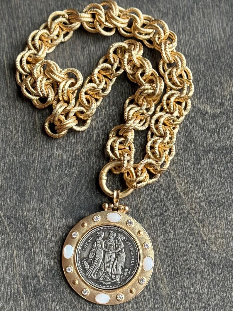 K&ouml;nnte beinhalten: Goldfarbene Halskette mit einem gro&szlig;en, kreisf&ouml;rmigen Anh&auml;nger, der ein detailliertes Relief von Figuren und die Inschrift "FOEDUS INVIOLABILE" aufweist. Der Anh&auml;nger ist mit klaren Kristallen und wei&szlig;en ovalen Einlagen verziert und h&auml;ngt an einer dicken, strukturierten Kette.