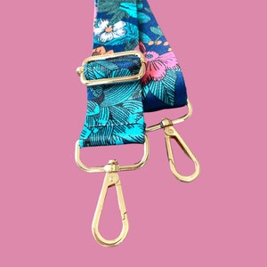 Pode incluir: Uma al&ccedil;a ajust&aacute;vel com fechos de metal dourado, com um padr&atilde;o floral azul e verde. A al&ccedil;a &eacute; feita de tecido e tem uma estampa floral.