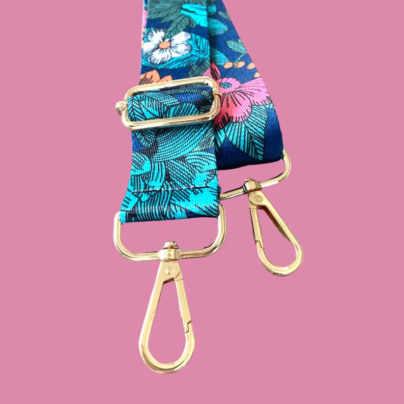 Pode incluir: Uma al&ccedil;a ajust&aacute;vel com fechos de metal dourado, com um padr&atilde;o floral azul e verde. A al&ccedil;a &eacute; feita de tecido e tem uma estampa floral.
