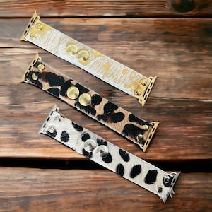 Op de afbeelding: Drie Apple Watch bandjes met verschillende dierenprintpatronen. De bandjes zijn gemaakt van leer en hebben gouden metalen accenten. De bandjes liggen op een houten oppervlak.