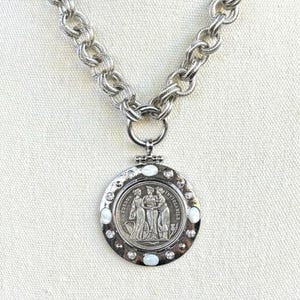 K&ouml;nnte beinhalten: Silberfarbene Halskette mit einem gro&szlig;en, runden Anh&auml;nger. Der Anh&auml;nger zeigt ein detailliertes Relief von drei Figuren und ist mit kleinen, wei&szlig;en Steinen verziert. Die Kette hat ein klobiges, strukturiertes Design.