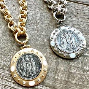 K&ouml;nnte beinhalten: Zwei Halsketten mit gro&szlig;en, klobigen Ketten. Eine ist goldfarben, die andere silberfarben. Jede hat einen runden Anh&auml;nger mit einem klassischen Reliefdesign, umgeben von Kristallen und Perlmuttakzenten. Die Anh&auml;nger tragen den Text "FOEDUS INVIOLABILE".