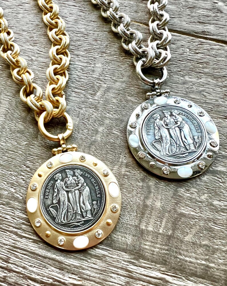 K&ouml;nnte beinhalten: Zwei Halsketten mit gro&szlig;en, klobigen Ketten. Eine ist goldfarben, die andere silberfarben. Jede hat einen runden Anh&auml;nger mit einem klassischen Reliefdesign, umgeben von Kristallen und Perlmuttakzenten. Die Anh&auml;nger tragen den Text "FOEDUS INVIOLABILE".